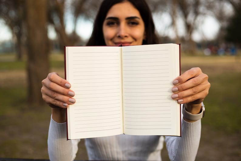 May include: A person holding a blank, lined notebook open with both hands. The notebook has a red cover and white pages.