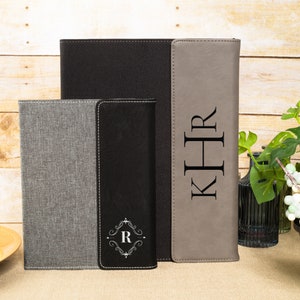 May include: Three personalized leatherette journals with different monogrammed initials. The journals are gray, black, and brown. The journals are on a wooden surface.