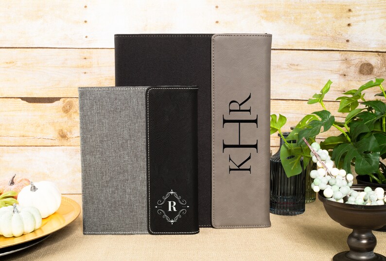May include: Three personalized leatherette journals with different monogrammed initials. The journals are gray, black, and brown. The journals are on a wooden surface.