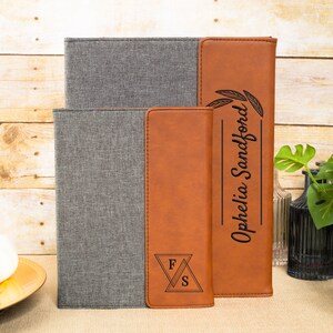 May include: Two personalized leather journals with a gray fabric cover. The larger journal has the name "Ophelia Sanford" engraved on the front with a floral design. The smaller journal has the initials "F.S." engraved on the front.