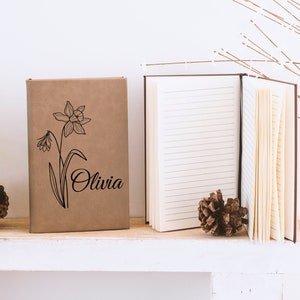 May include: A brown notebook with a black line drawing of a daffodil and the name "Olivia" on the cover. The notebook is sitting on a white shelf with a pine cone on either side.