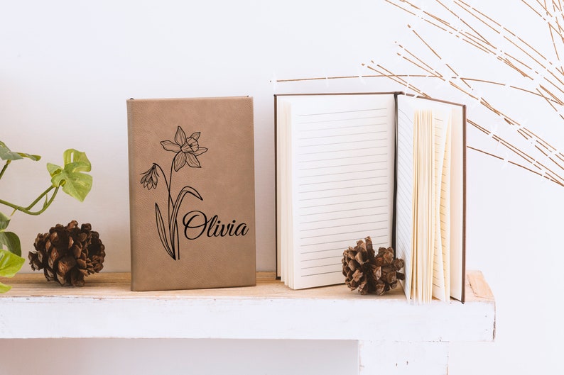 May include: A brown notebook with a black line drawing of a daffodil and the name "Olivia" on the cover. The notebook is sitting on a white shelf with a pine cone on either side.