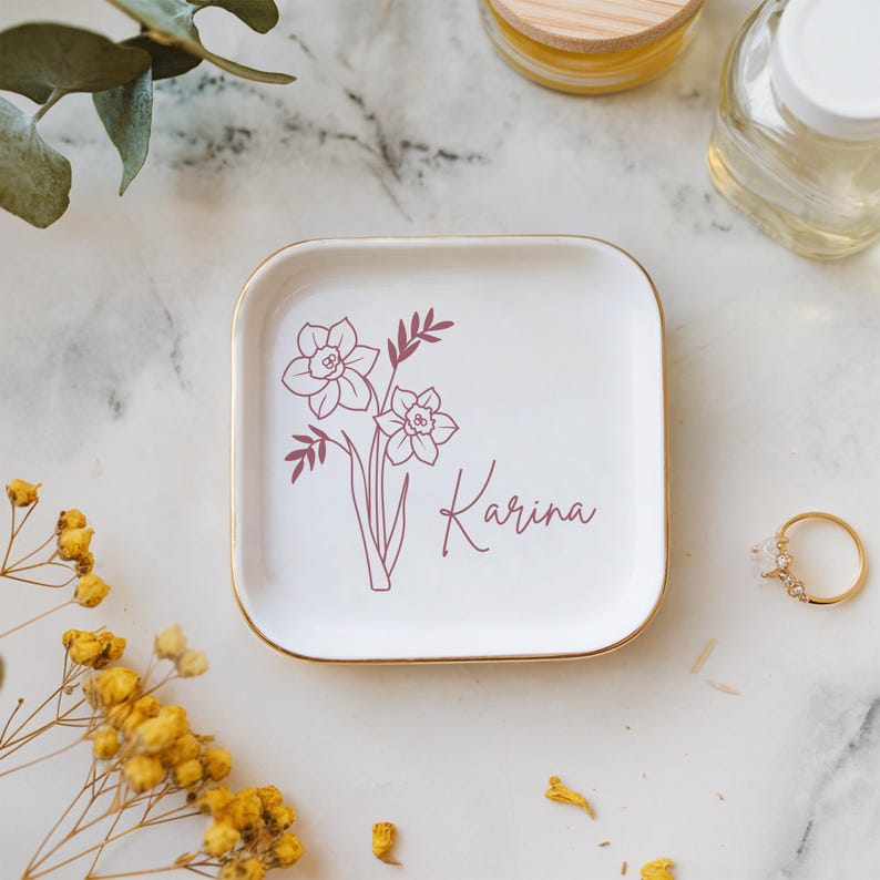 Op de afbeelding: Witte keramische sieradenbakje met een gouden rand. Het bakje heeft een roze lijntekening van een boeket narcissen en de naam "Karina" in cursief.