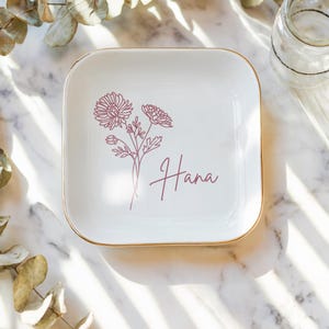 Op de afbeelding: Witte keramische schaal met een gouden rand, versierd met een roze lijntekening van bloemen en de naam "Hana" in roze schrift.