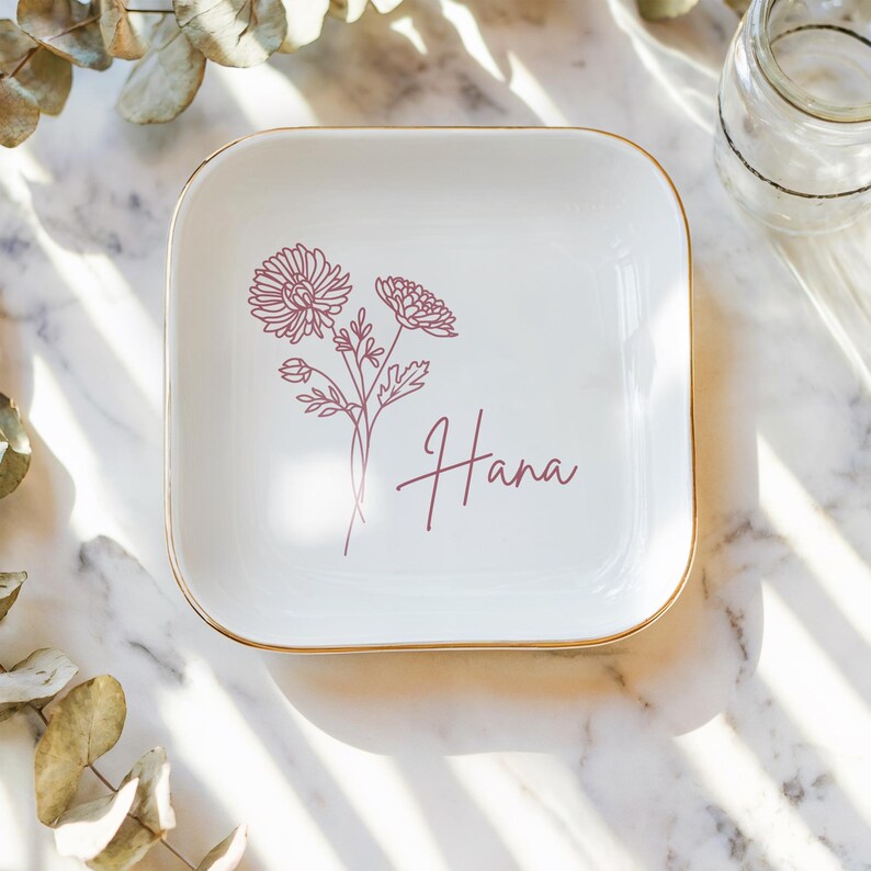 Op de afbeelding: Witte keramische schaal met een gouden rand, versierd met een roze lijntekening van bloemen en de naam "Hana" in roze schrift.
