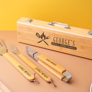 May include: A wooden barbecue tool set with a spatula, fork, knife, and tongs, all with wooden handles. The set includes a wooden box with the text "GEORGE'S GRILL AND CHILL". Each tool is engraved with "GEORGE MENDES".