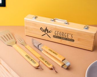 May include: A wooden barbecue tool set with a spatula, fork, knife, and tongs, all with wooden handles. The set includes a wooden box with the text "GEORGE'S GRILL AND CHILL". Each tool is engraved with "GEORGE MENDES".