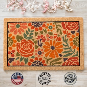 May include: A coir doormat with a floral design featuring orange, red, and green flowers. The doormat has a black border and is made in the USA.