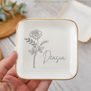 Op de afbeelding: Witte vierkante sieradenbak met een gouden rand. De bak heeft een grijze lijntekening van een roos met de naam "Deasia" eronder.