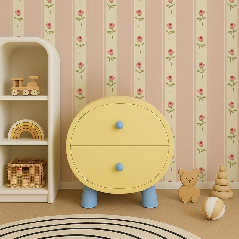 May include: A yellow, round two-drawer cabinet with blue knobs and legs. A white shelf holds a wooden train, rainbow, and basket. A wooden bear, stacking rings, and ball sit on the floor. The wallpaper has pink stripes and floral patterns.