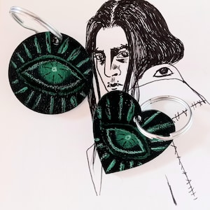 May include: Two leather keychains with green eye designs. One keychain is round and the other is heart-shaped.