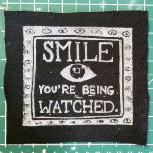 Pode incluir: Remendo de tecido preto com uma borda branca e texto branco que diz "SMILE YOU'RE BEING WATCHED." Dentro da borda há um olho branco com uma pupila preta.