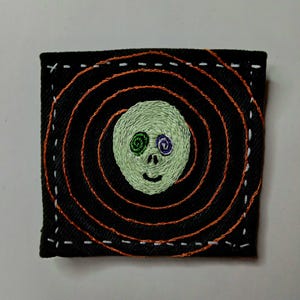 Op de afbeelding: Gestikt patch met een gezicht met groene en paarse ogen op een zwarte achtergrond met oranje en witte stiksels.