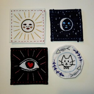 May include: Four embroidered patches featuring different designs. The first patch is white with a yellow sun with a red heart and a smiling face. The second patch is blue denim with a white crescent moon with a blue face. The third patch is black with a white eye with a red heart. The fourth patch is white with a blue cat with a bow tie and a blue fishbone design around the edge.