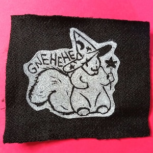 May include: A black fabric patch with a white embroidered image of a squirrel wearing a witch's hat and holding a flower. The text "GNEHEHEHA" is embroidered above the squirrel.