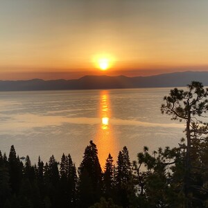 Pode incluir: Um p&ocirc;r do sol c&ecirc;nico sobre um lago com montanhas ao fundo. O sol se p&otilde;e atr&aacute;s das montanhas, lan&ccedil;ando um brilho dourado sobre a &aacute;gua. H&aacute; &aacute;rvores em primeiro plano, silhuetas contra o c&eacute;u.