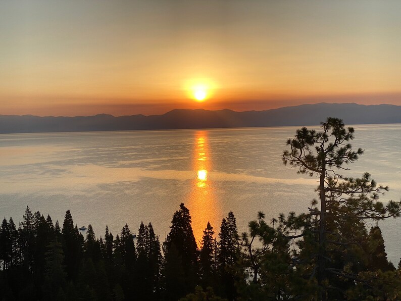 Pode incluir: Um p&ocirc;r do sol c&ecirc;nico sobre um lago com montanhas ao fundo. O sol se p&otilde;e atr&aacute;s das montanhas, lan&ccedil;ando um brilho dourado sobre a &aacute;gua. H&aacute; &aacute;rvores em primeiro plano, silhuetas contra o c&eacute;u.