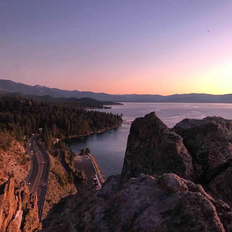 Pode incluir: Uma vista panor&acirc;mica de um penhasco rochoso com vista para uma estrada sinuosa que leva a um lago. O c&eacute;u &eacute; de um roxo e rosa suaves, criando um belo cen&aacute;rio de p&ocirc;r do sol.