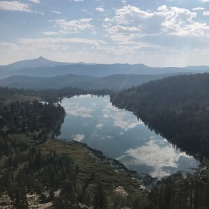 Pode incluir: Uma vista do topo de uma montanha com vista para um lago cercado por &aacute;rvores. O lago reflete o c&eacute;u azul e as nuvens brancas.