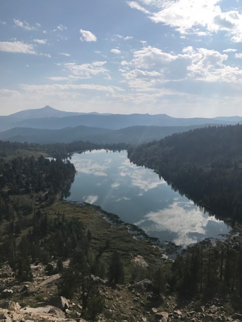 Pode incluir: Uma vista do topo de uma montanha com vista para um lago cercado por &aacute;rvores. O lago reflete o c&eacute;u azul e as nuvens brancas.