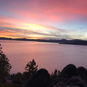 Pode incluir: Uma vista panor&acirc;mica de um lago com um c&eacute;u de p&ocirc;r do sol rosa e laranja. O lago est&aacute; coberto de nevoeiro e as montanhas ao longe est&atilde;o silhuetas contra o c&eacute;u. Um par de p&eacute;s est&aacute; em primeiro plano, descansando sobre uma rocha.