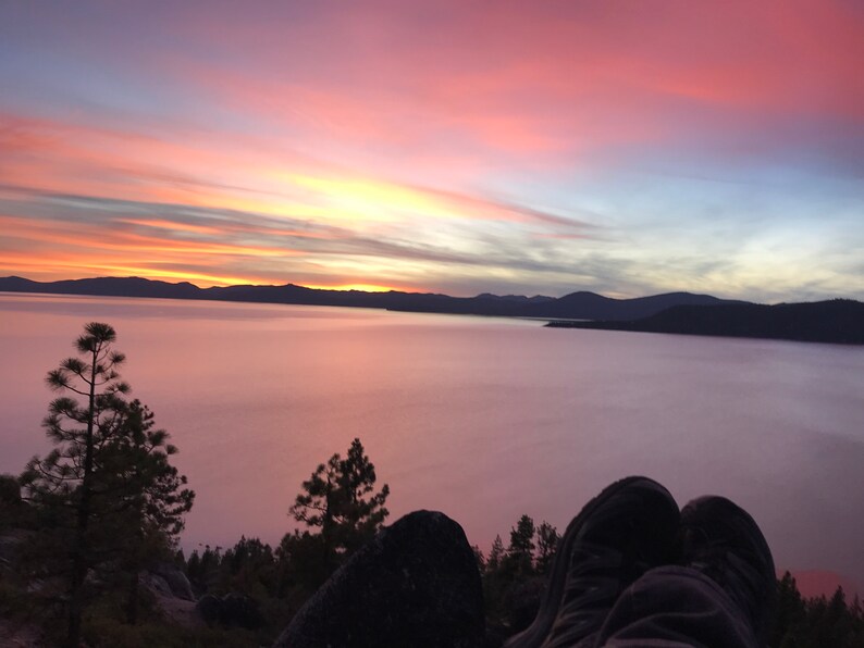 Pode incluir: Uma vista panor&acirc;mica de um lago com um c&eacute;u de p&ocirc;r do sol rosa e laranja. O lago est&aacute; coberto de nevoeiro e as montanhas ao longe est&atilde;o silhuetas contra o c&eacute;u. Um par de p&eacute;s est&aacute; em primeiro plano, descansando sobre uma rocha.
