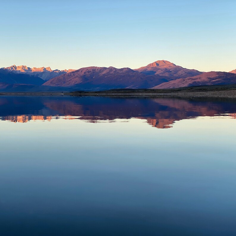 Pode incluir: Uma cordilheira de montanhas refletida em um lago tranquilo ao nascer do sol. O c&eacute;u &eacute; azul claro e as montanhas s&atilde;o rosa e roxo suaves.
