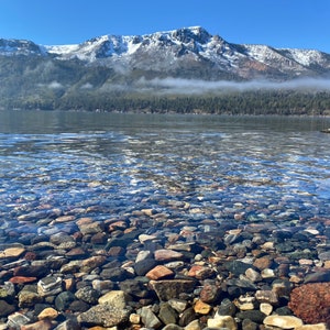 Pode incluir: Um lago de montanha claro com um fundo rochoso e uma cordilheira nevada ao fundo.