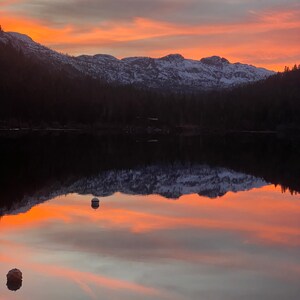 Pode incluir: Uma vista panor&acirc;mica de um lago que reflete uma cordilheira ao p&ocirc;r do sol. O c&eacute;u &eacute; de um laranja e rosa vibrantes, com nuvens que projetam longas sombras sobre a &aacute;gua.