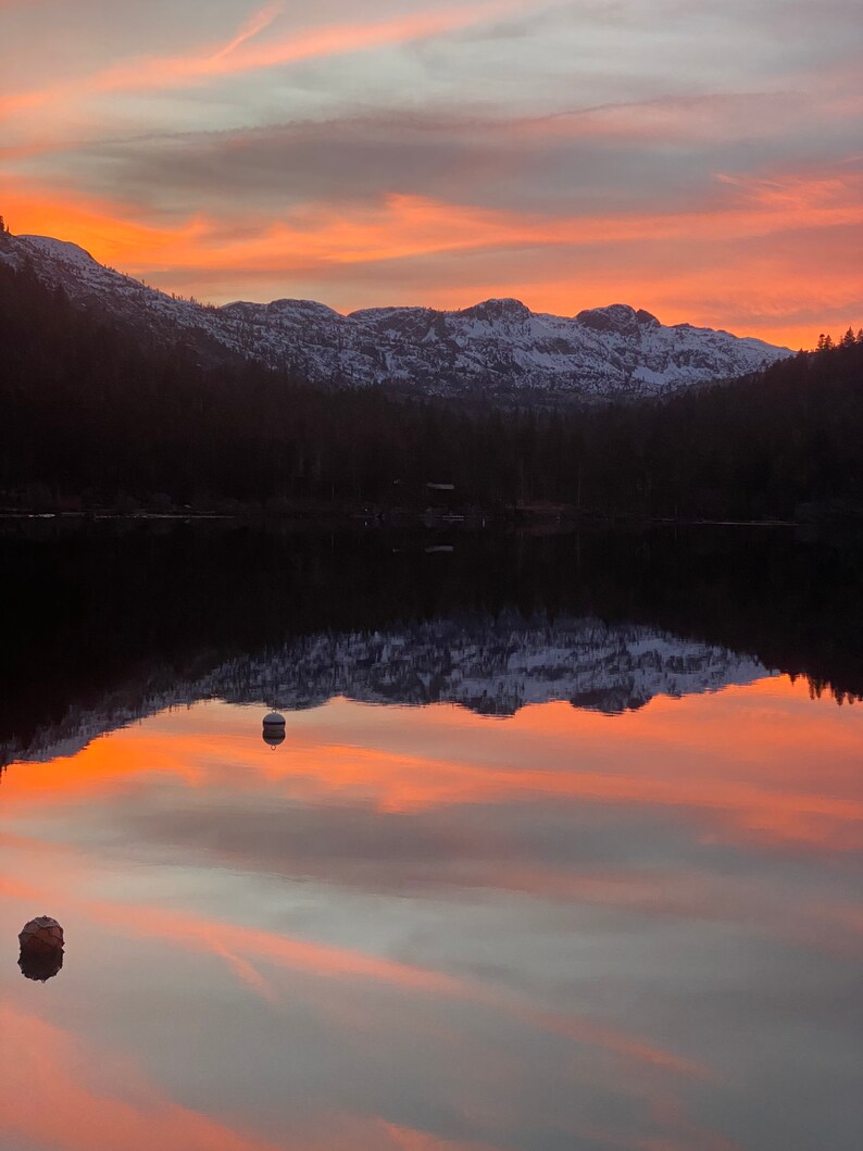 Pode incluir: Uma vista panor&acirc;mica de um lago que reflete uma cordilheira ao p&ocirc;r do sol. O c&eacute;u &eacute; de um laranja e rosa vibrantes, com nuvens que projetam longas sombras sobre a &aacute;gua.