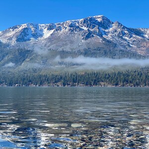 Pode incluir: Uma cordilheira com picos cobertos de neve e uma camada de nevoeiro no vale, refletida na &aacute;gua calma de um lago.