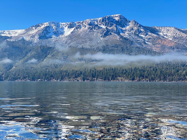 Pode incluir: Uma cordilheira com picos cobertos de neve e uma camada de nevoeiro no vale, refletida na &aacute;gua calma de um lago.