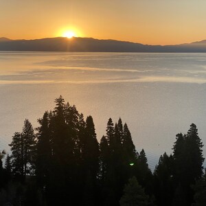 Pode incluir: Um nascer do sol c&ecirc;nico sobre um lago com uma cordilheira de montanhas ao fundo. O sol nasce sobre as montanhas, lan&ccedil;ando um brilho dourado sobre a &aacute;gua. Silhuetas de &aacute;rvores margeiam a costa em primeiro plano.