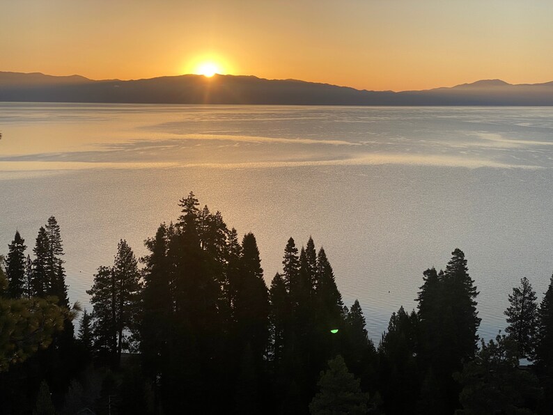 Pode incluir: Um nascer do sol c&ecirc;nico sobre um lago com uma cordilheira de montanhas ao fundo. O sol nasce sobre as montanhas, lan&ccedil;ando um brilho dourado sobre a &aacute;gua. Silhuetas de &aacute;rvores margeiam a costa em primeiro plano.