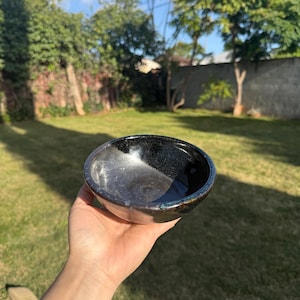 May include: A small, round, black ceramic bowl with a glossy finish. The bowl has a dark interior and a slightly iridescent rim. The bowl is held in a hand against a backdrop of green grass and trees.