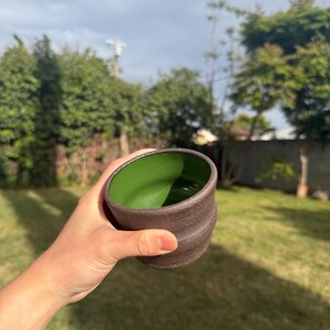 Peut inclure: Une tasse en céramique faite à la main avec un extérieur brun foncé et un intérieur vert vif. La tasse a un design texturé et strié et est tenue sur fond d'herbe verte et de ciel nuageux. La tasse mesure environ 10 cm de hauteur.