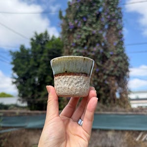 May include: A small, handmade ceramic cup with a textured, earthy design. The top portion is a muted green, transitioning to a brown, textured middle, and a speckled, light brown base. The cup is held in a hand against a blurred outdoor background.