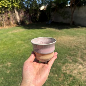 May include: A small ceramic cup featuring a smooth, rounded shape with a two-tone glaze. The upper portion is finished in a soft, dusty pink, while the lower section transitions into a natural, unglazed beige clay base with a visible horizontal line.