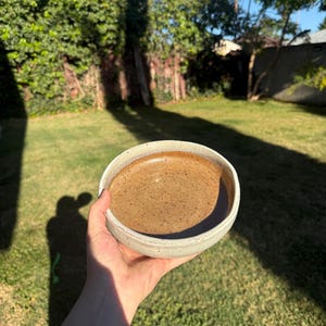May include: A handcrafted ceramic bowl with a pale grey exterior and a brown interior. The bowl is held in a hand against a backdrop of green grass and trees, with sunlight casting shadows.