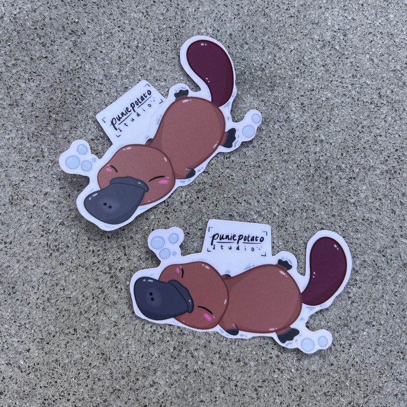 May include: Two brown and white cartoon platypus stickers with the text "puni potato studio" on each. The platypus are swimming with bubbles around them.