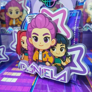 Może przedstawiać: Wielowarstwowa papierowa dekoracja przedstawiająca trzy dziewczyny z kreskówek z fioletowymi, czerwonymi i czarnymi włosami. Centralna postać ma fioletowy warkocz i nosi żółtą kurtkę. Nazwa "DANIELA" jest wydrukowana na białym tle.