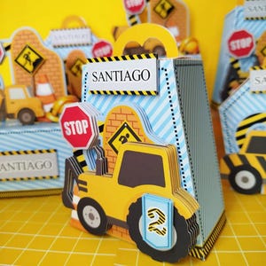 May include: A construction-themed gift bag with a yellow tractor, stop sign, and the name "SANTIAGO". The bag has blue and white stripes and a yellow handle. Other similar bags are in the background.