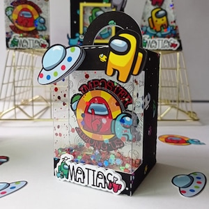 Puede incluir: Una caja de regalo de fiesta en blanco y negro con una ventana transparente. La caja tiene un asa y está decorada con personajes de dibujos animados coloridos del videojuego Among Us. La caja está llena de confeti de colores y tiene las palabras "IMPOSTER AMONG US" y "MATIAS" impresas en ella.