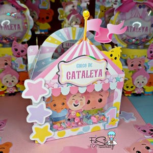 Op de afbeelding: Een roze en gele kartonnen doos in de vorm van een circus tent met een doorzichtig venster. De doos heeft een handvat en er staat "Circo de Cataleya" op de voorkant. Er zitten drie cartoon varkensfiguren in het venster.