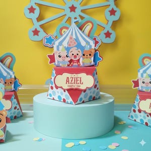 Pode incluir: Decoração festiva de aniversário com tema de circo. A caixa apresenta o nome "AZIEL" e "CUMPLE UNO". É adornada com personagens de desenhos animados e um fundo de roda gigante. A combinação de cores inclui vermelho, azul e branco.
