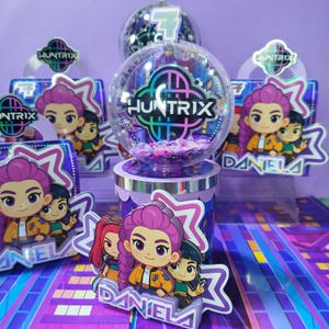 Puede incluir: Un conjunto de decoración para fiestas de cumpleaños con un globo transparente que muestra "HUNTRIX" y confeti. El conjunto incluye recortes de personajes, adornos para pasteles y una combinación de colores púrpura y plateado. También es visible el número "7".
