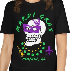 May include: Black t-shirt with a Mardi Gras design. The graphic features a white skull wearing a purple mask and a green and gold beaded necklace. The words "Mardi Gras" and "Mobile, AL" are in green.