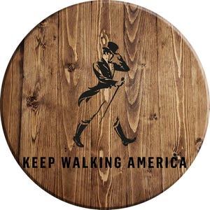 Puede incluir: Un letrero redondo de madera con un acabado de grano de madera marr&oacute;n. El letrero presenta una silueta negra de un hombre caminando con un bast&oacute;n y el texto "KEEP WALKING AMERICA".
