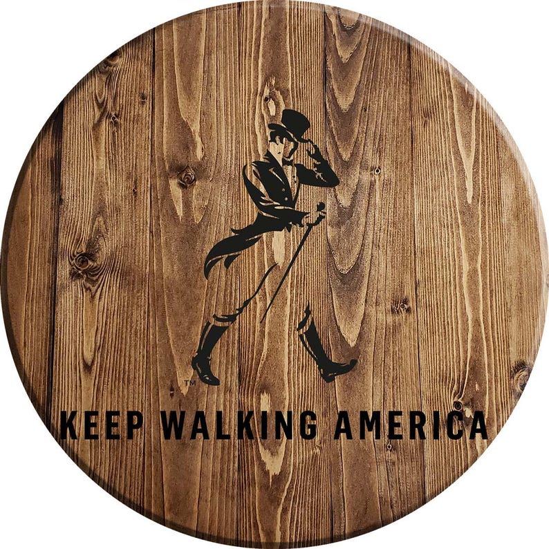 Puede incluir: Un letrero redondo de madera con un acabado de grano de madera marr&oacute;n. El letrero presenta una silueta negra de un hombre caminando con un bast&oacute;n y el texto "KEEP WALKING AMERICA".