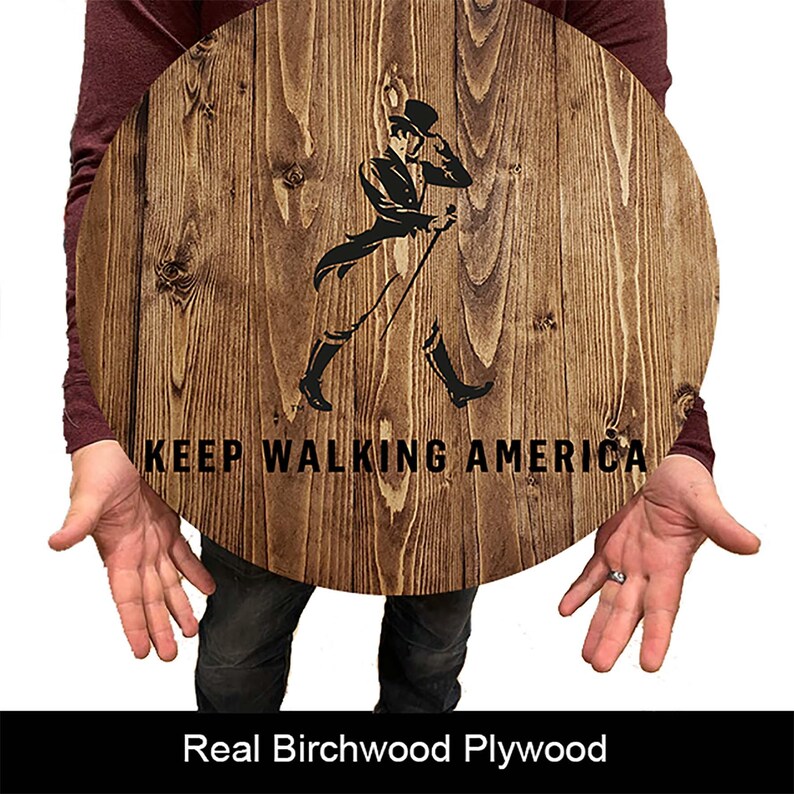 Puede incluir: Un letrero redondo de madera con un acabado de grano de madera marr&oacute;n. El letrero presenta una silueta negra de un hombre con un sombrero de copa caminando con un bast&oacute;n. El texto "KEEP WALKING AMERICA" est&aacute; impreso en negro debajo de la silueta.